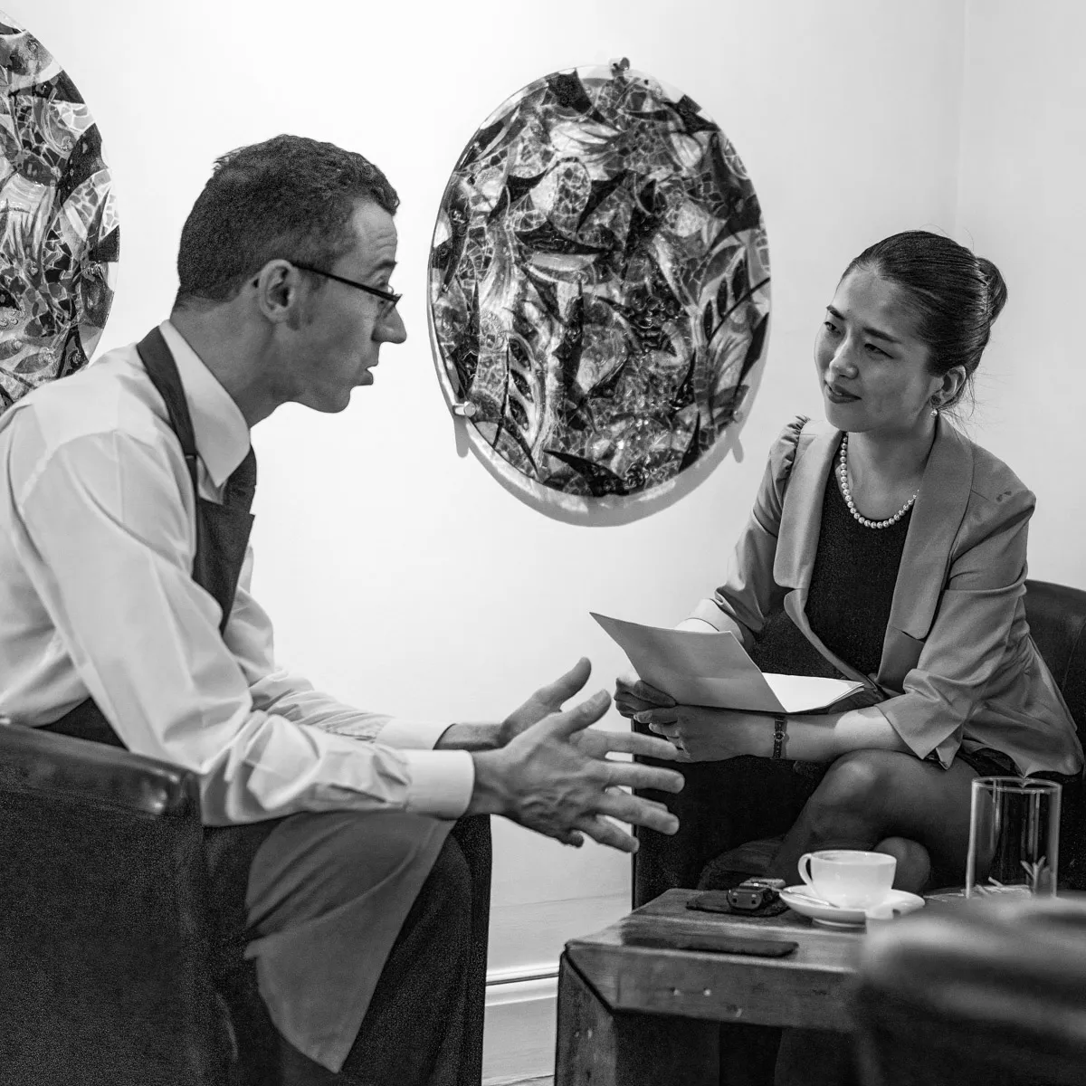 Black and white candid photograph of the sommelier gesturing while in conversation with a guest in a leather armchair at Sat Bains