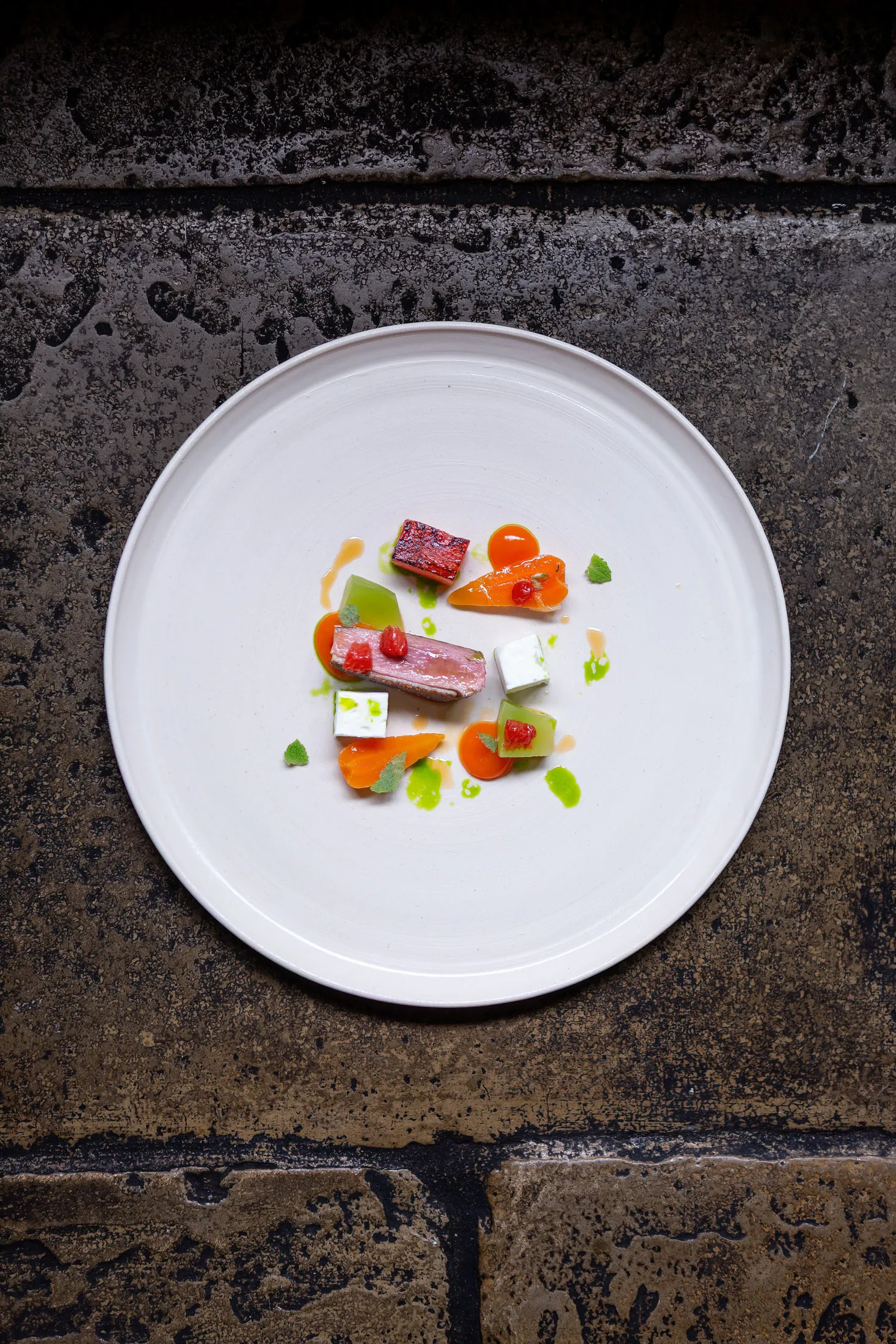 Top-down view of a savory main course at Sat Bains, medium-rare meat with geometric garnishes, orange puree dots and green oil on a round white plate