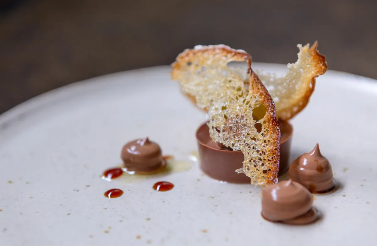 A smooth dark chocolate disc on a white plate, topped with delicate translucent golden sugar glass shards at Sat Bains