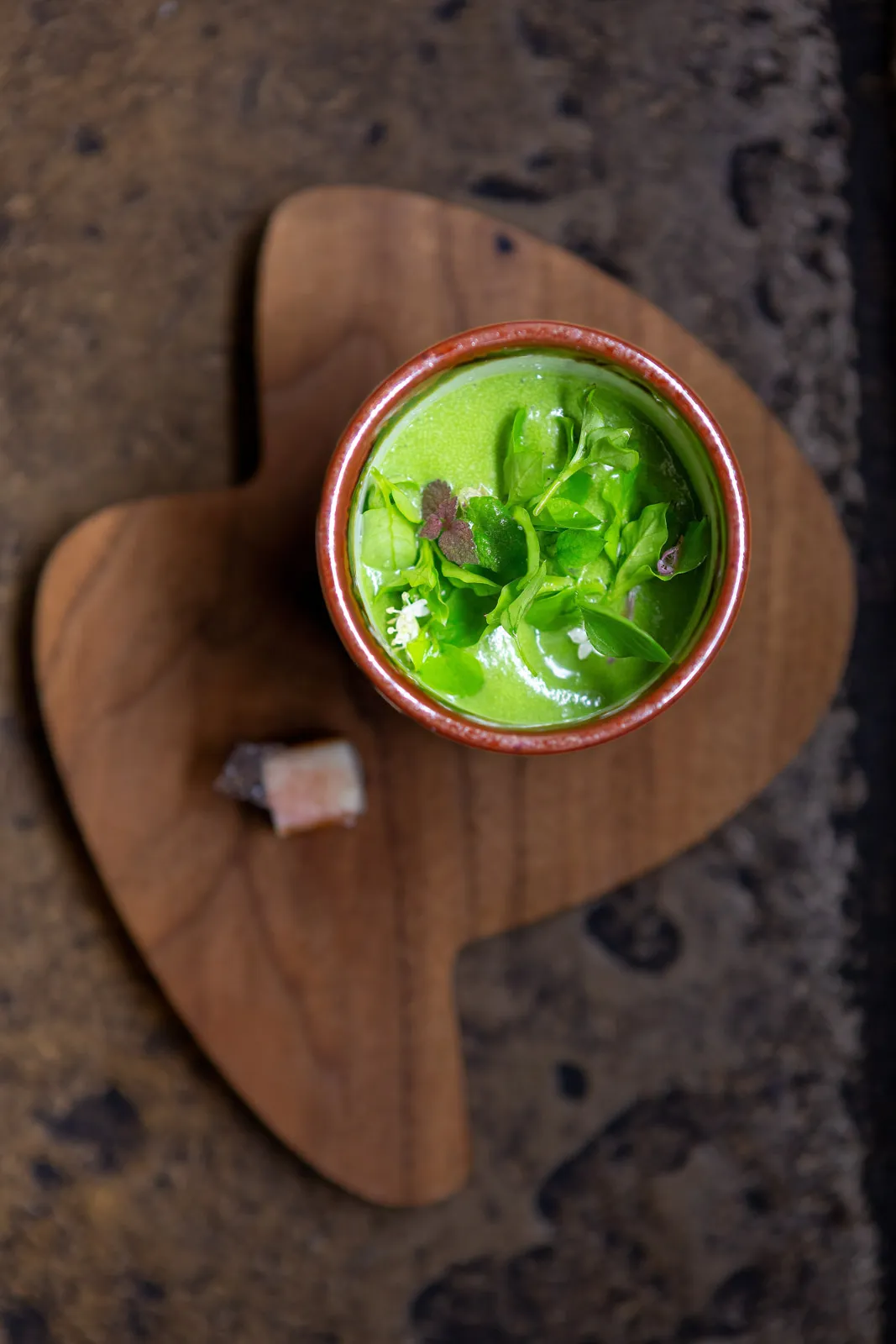 Vibrant green soup or puree in a small dark ceramic bowl, garnished with microgreens and white blossoms on an organic wooden board
