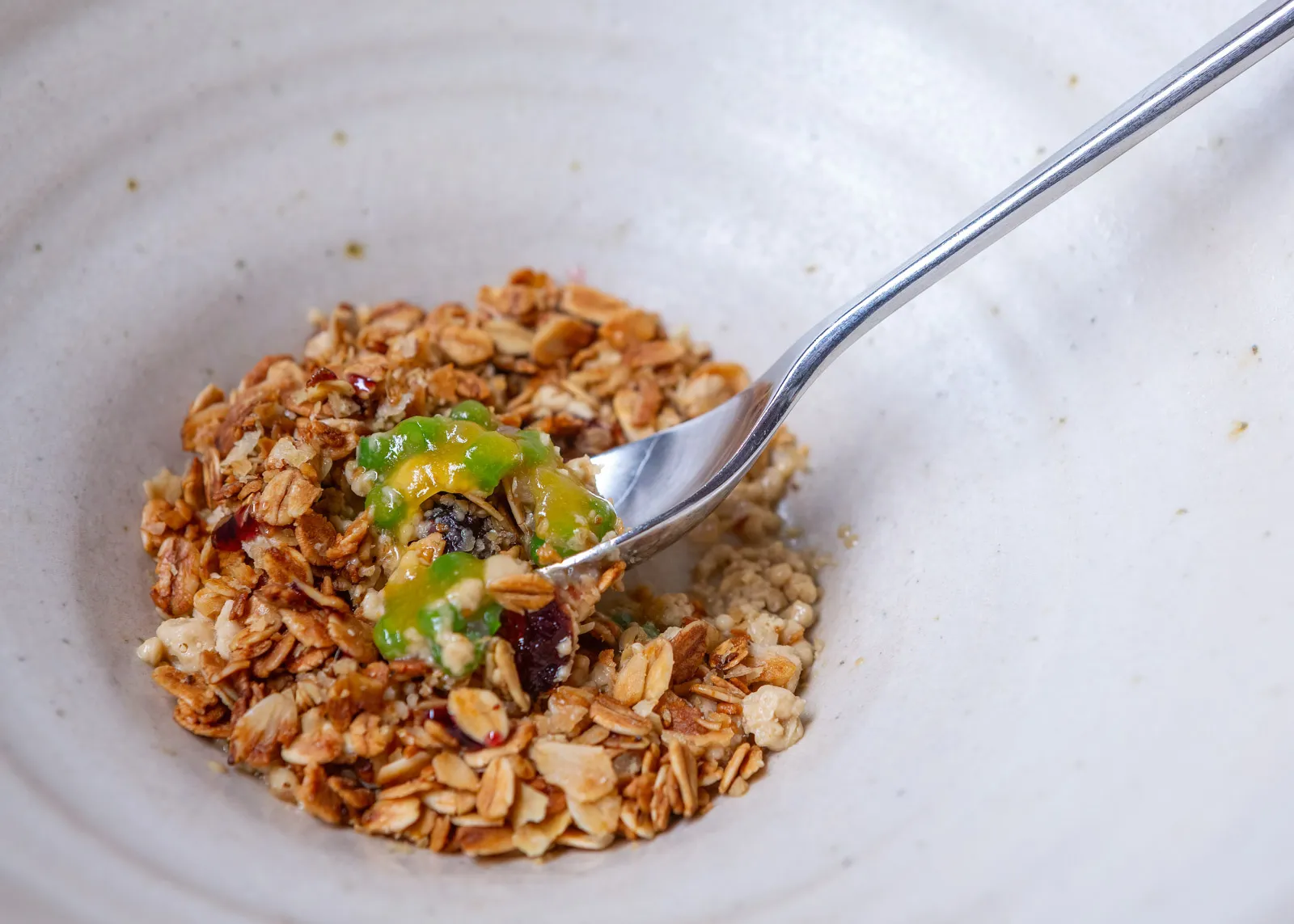 A breakfast-inspired course in a ceramic bowl, granola, oats and nuts with vibrant green and yellow fruit gel, a silver spoon lifting a bite
