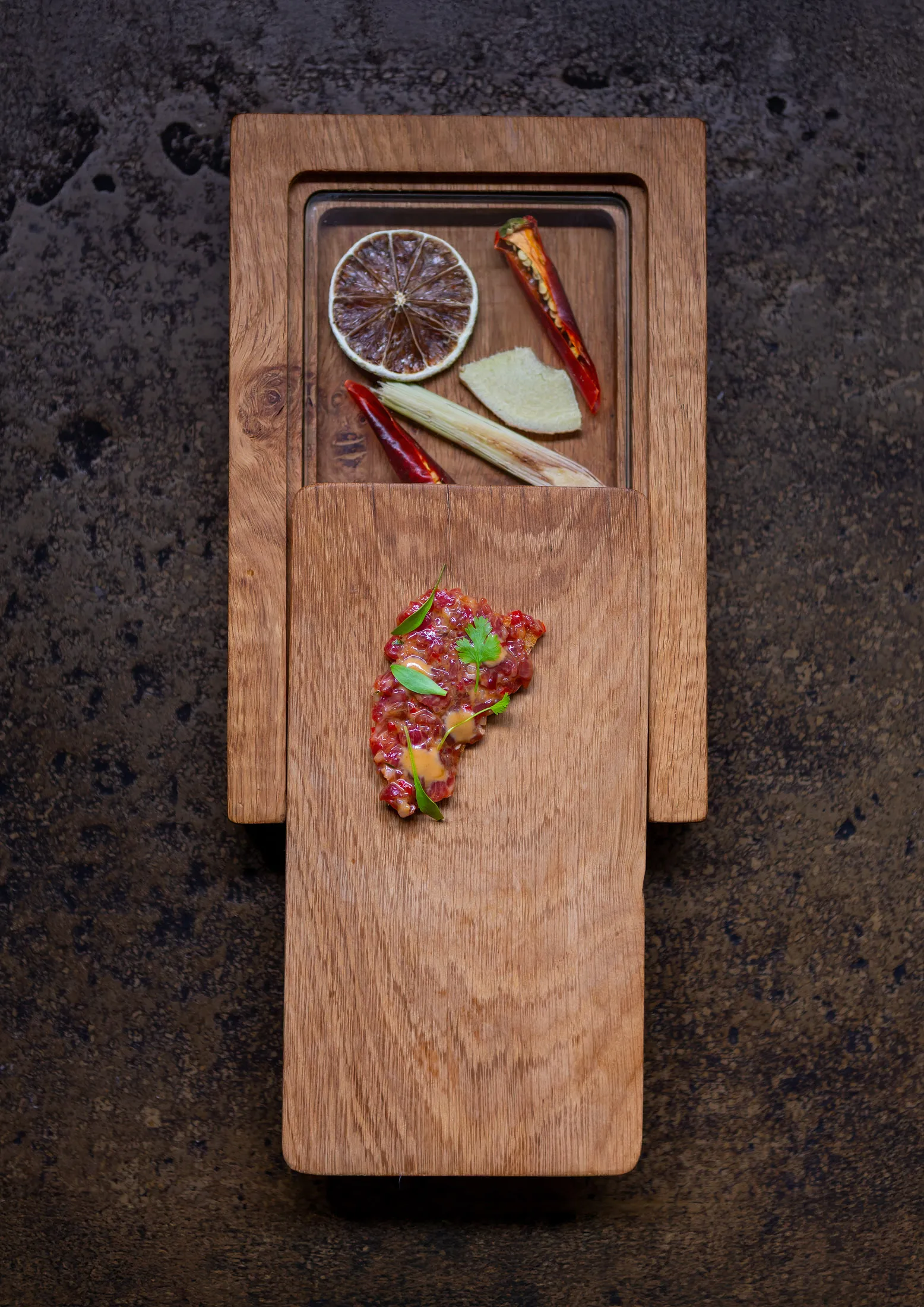 Beef tartare with microgreens on a sliding wooden box structure that opens to reveal aromatics, lemongrass, dried citrus and chili