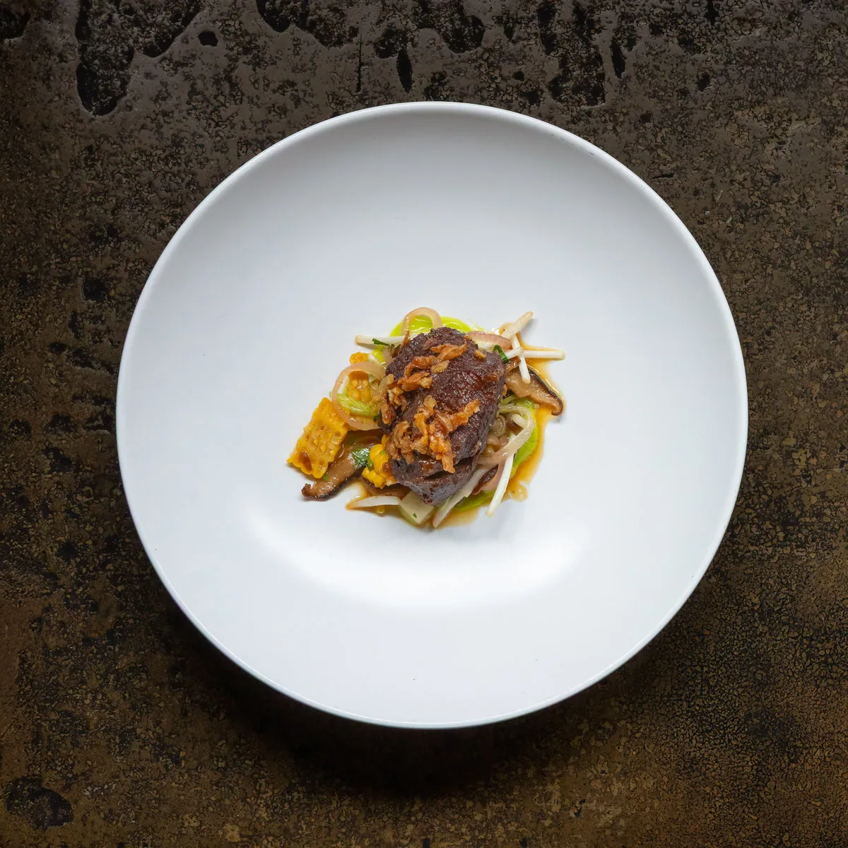 Glazed beef with crispy fried onions, sweetcorn, mushrooms and green herb oil in a wide white bowl at Restaurant Sat Bains