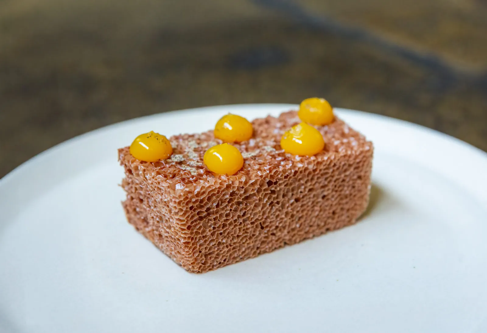 A rectangular block of aerated chocolate sponge on a white plate, topped with five glossy yellow gel spheres and a dusting of spice