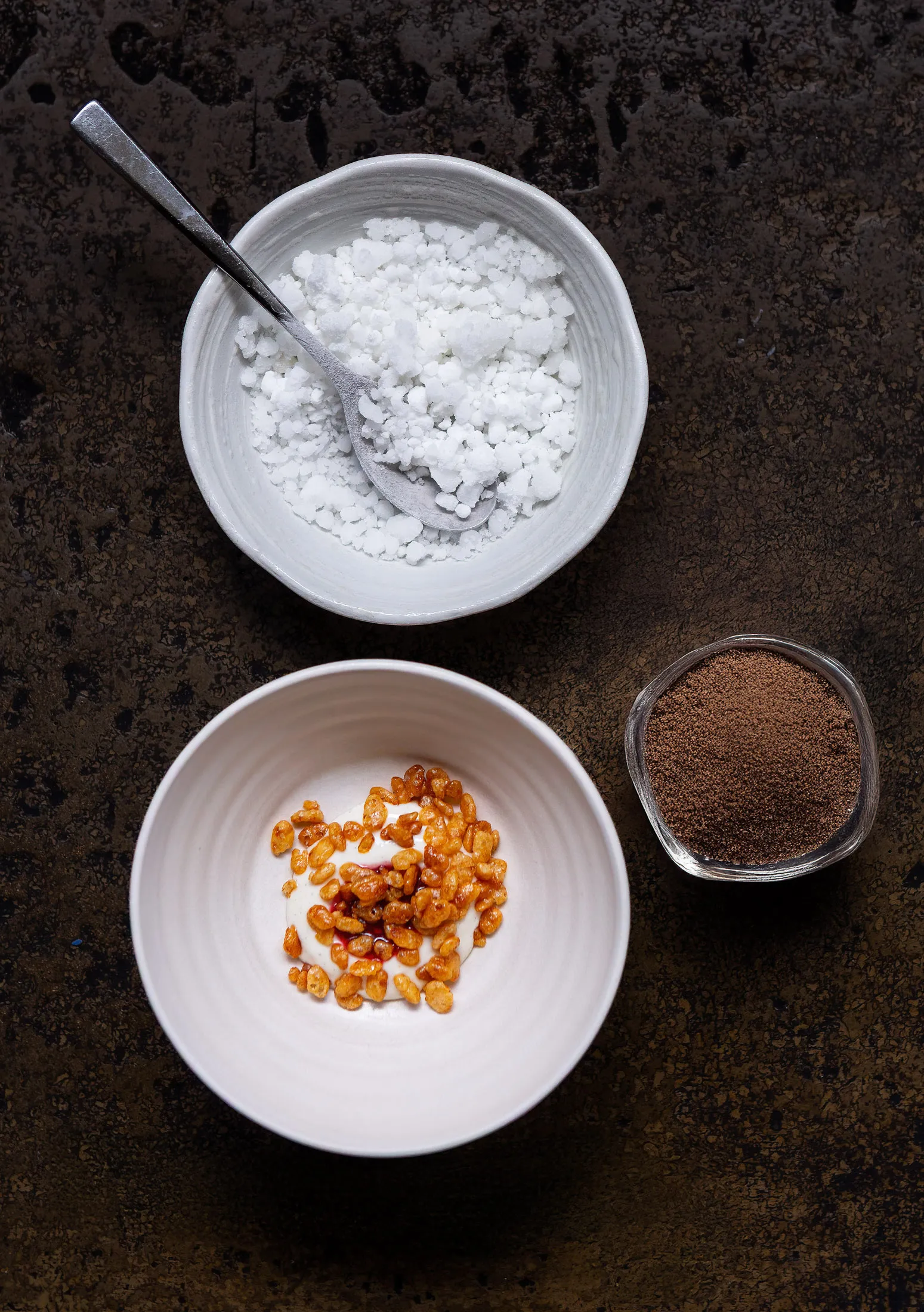 Top-down dessert layout at Sat Bains, a creamy bowl with caramelised grains and red syrup swirl, a bowl of white granita crystals, a dish of brown praline powder