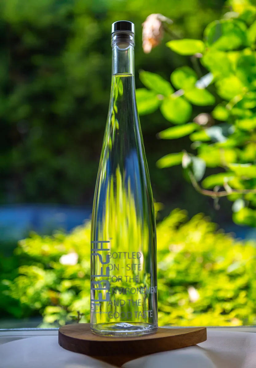 A tall clear glass water bottle bottled on-site at Restaurant Sat Bains, Nottingham, a brightly lit garden visible behind