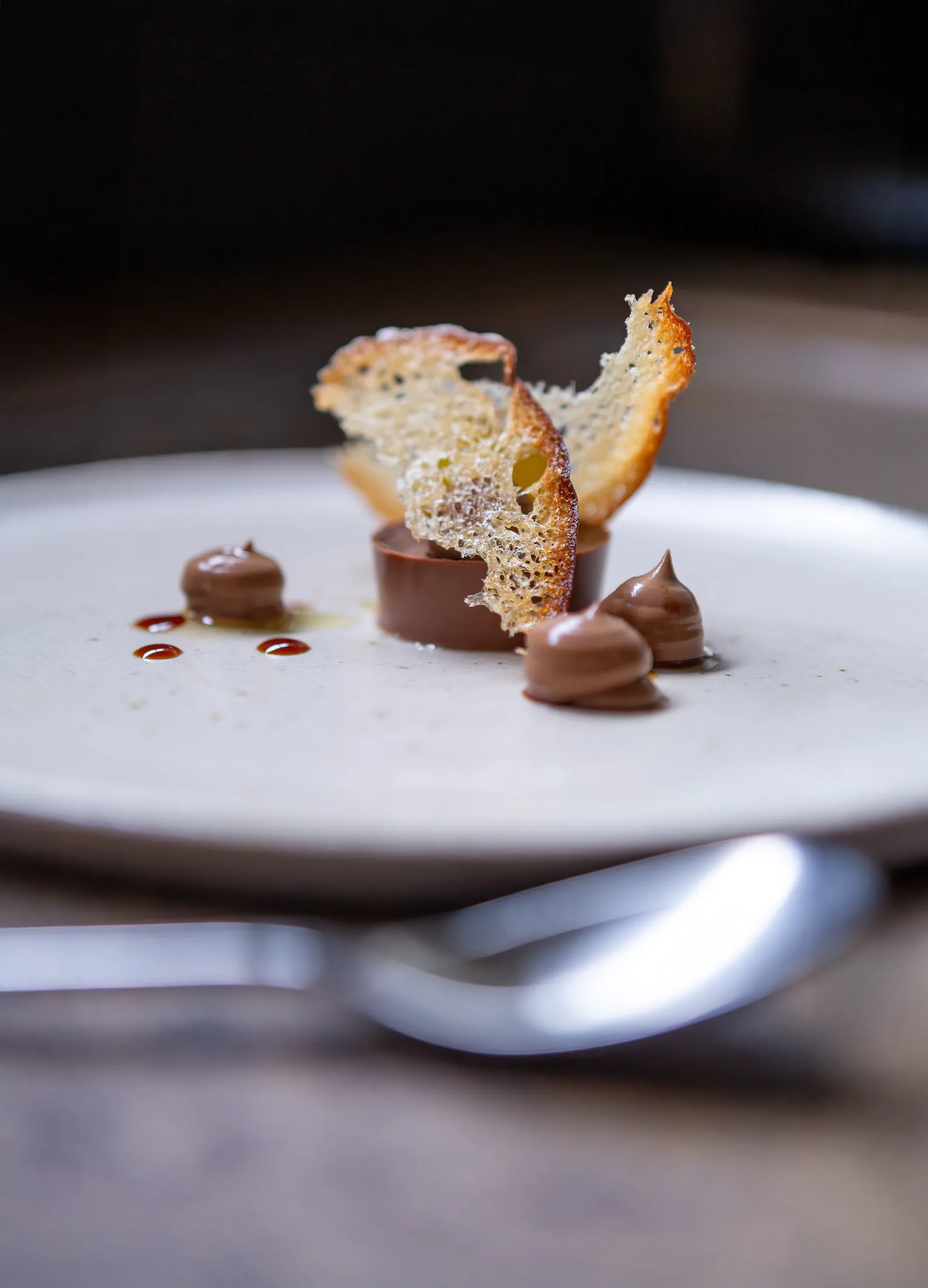 Chocolate ganache disc on a white plate with two upright lacy golden tuile shards, drops of chocolate cream and dark syrup at Sat Bains