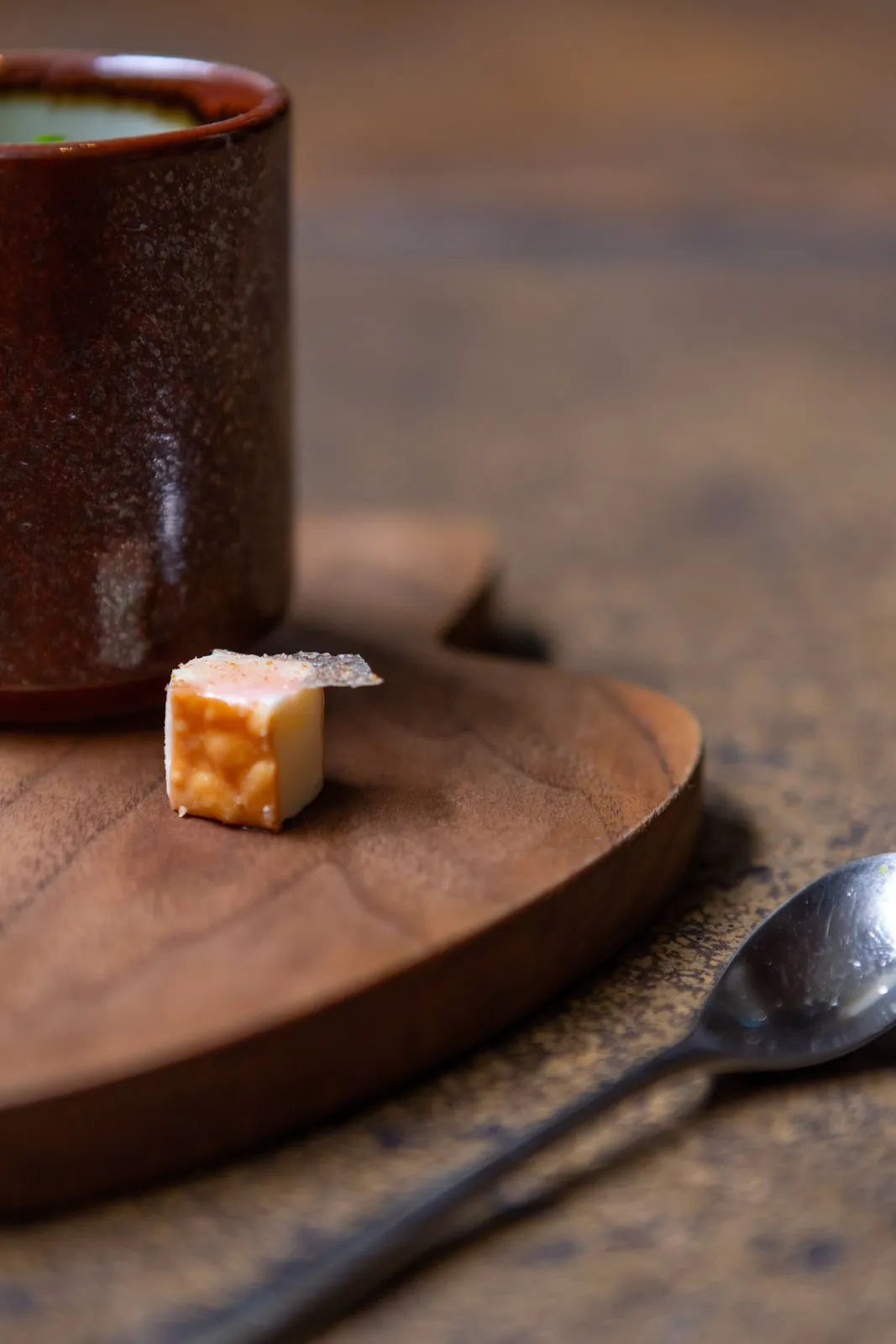 A tiny cube of salmon confection on an organic wooden board, a textured dark ceramic cup softly out of focus in the background