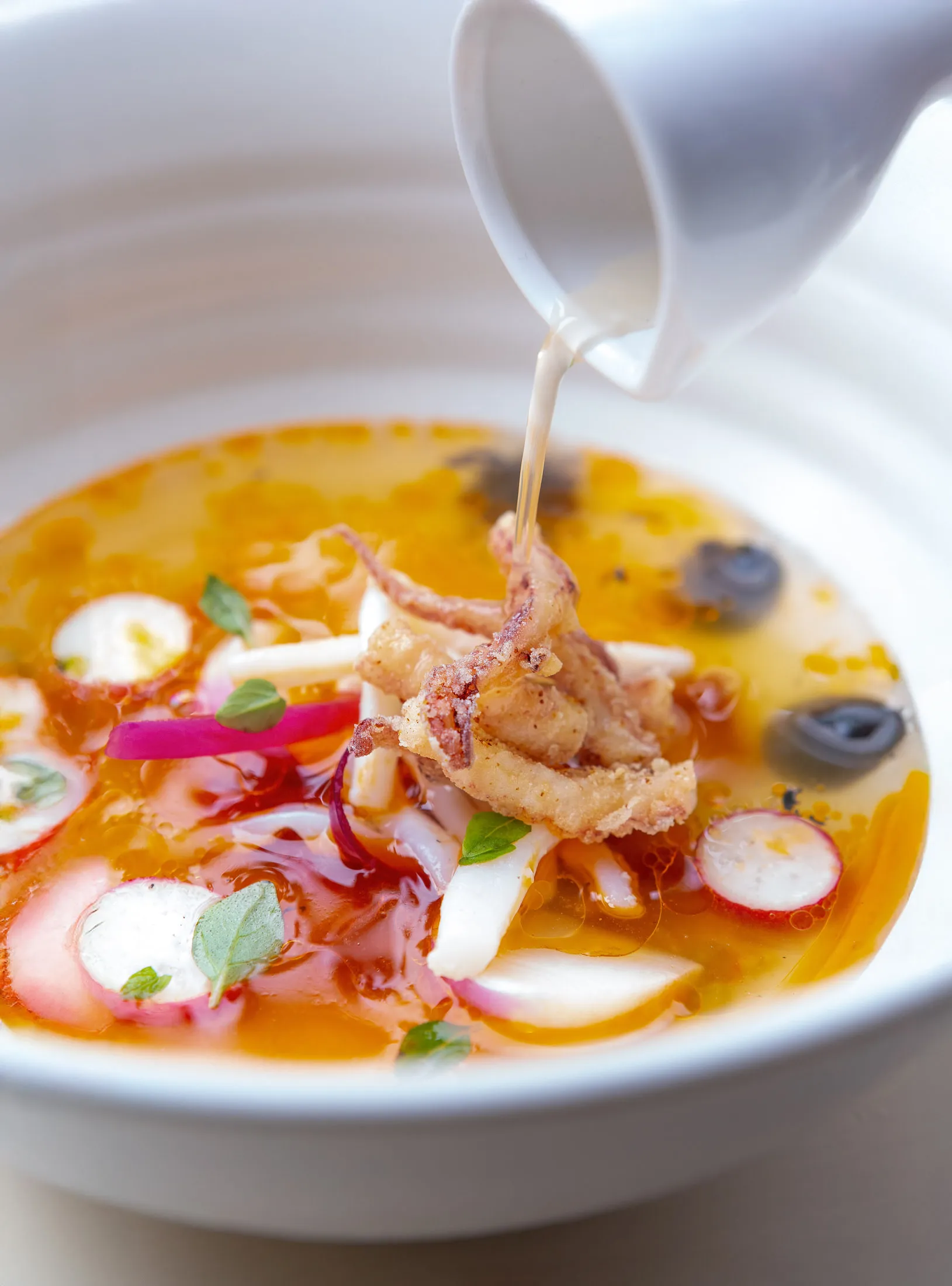 Squid dish being finished tableside, clear broth poured from a white ceramic pitcher into a bowl of fried squid, radishes and orange oil
