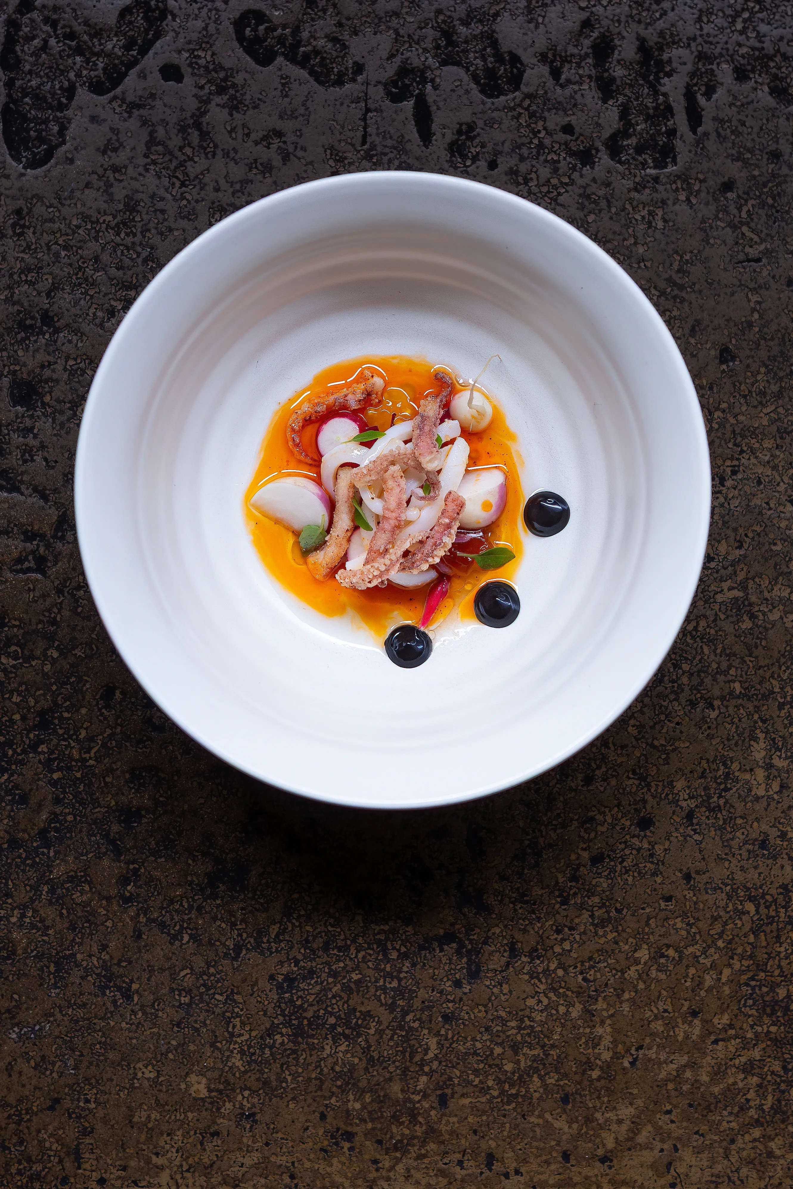 Overhead view of the squid dish in a white bowl against dark textured stone at Restaurant Sat Bains