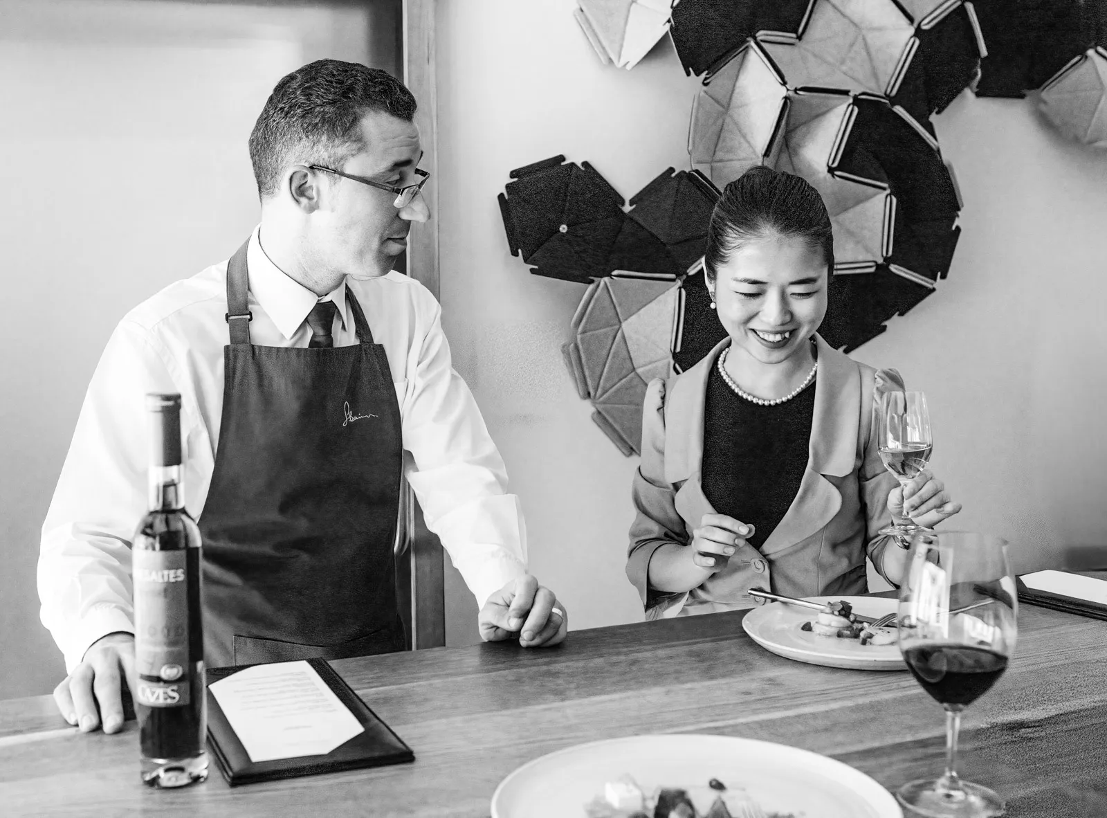 Black and white candid at the chef's table at Sat Bains, the sommelier gesturing while speaking to a guest examining a wine glass