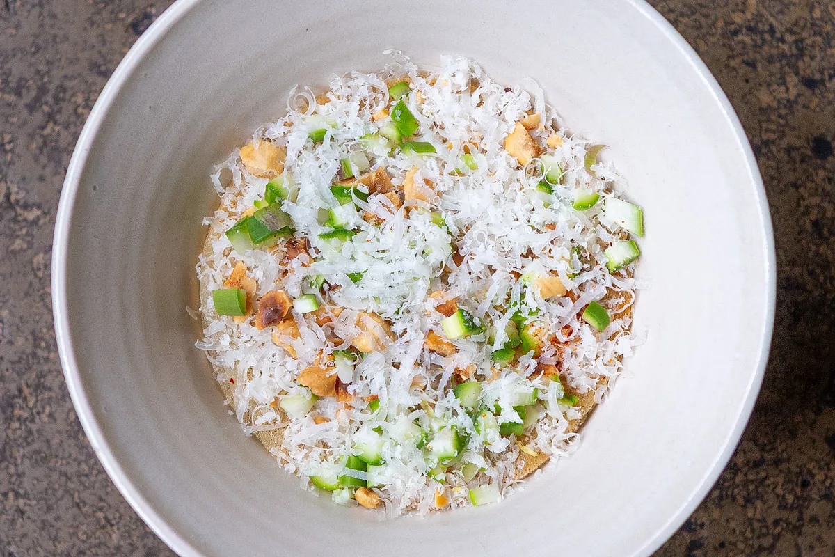 Top-down close-up of a white bowl with a snow-like layer of grated parmesan, diced green vegetables and toasted grains