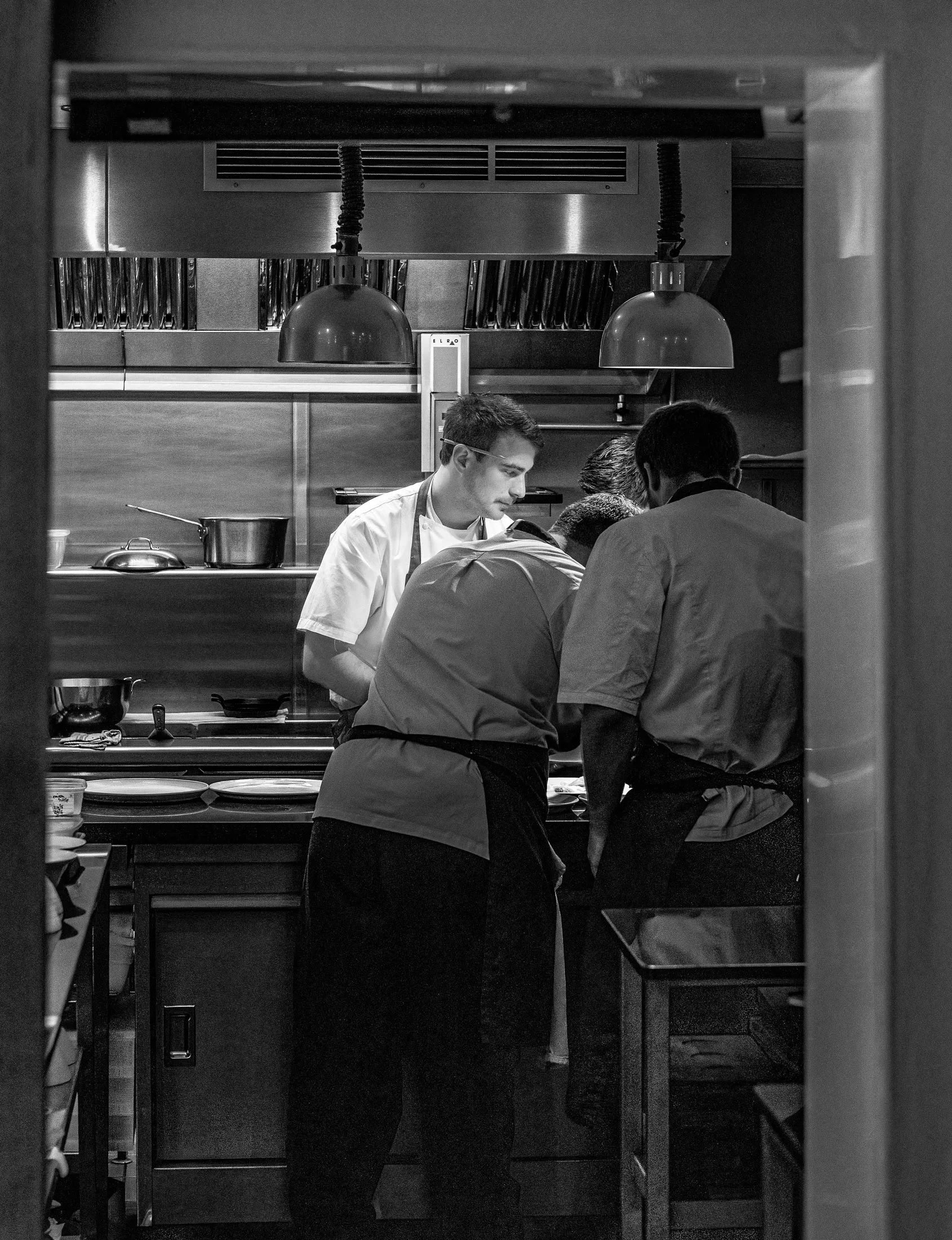 Kitchen team at Restaurant Sat Bains huddled over the pass under heat lamps, intensely focused on finishing dishes, the full kitchen visible behind
